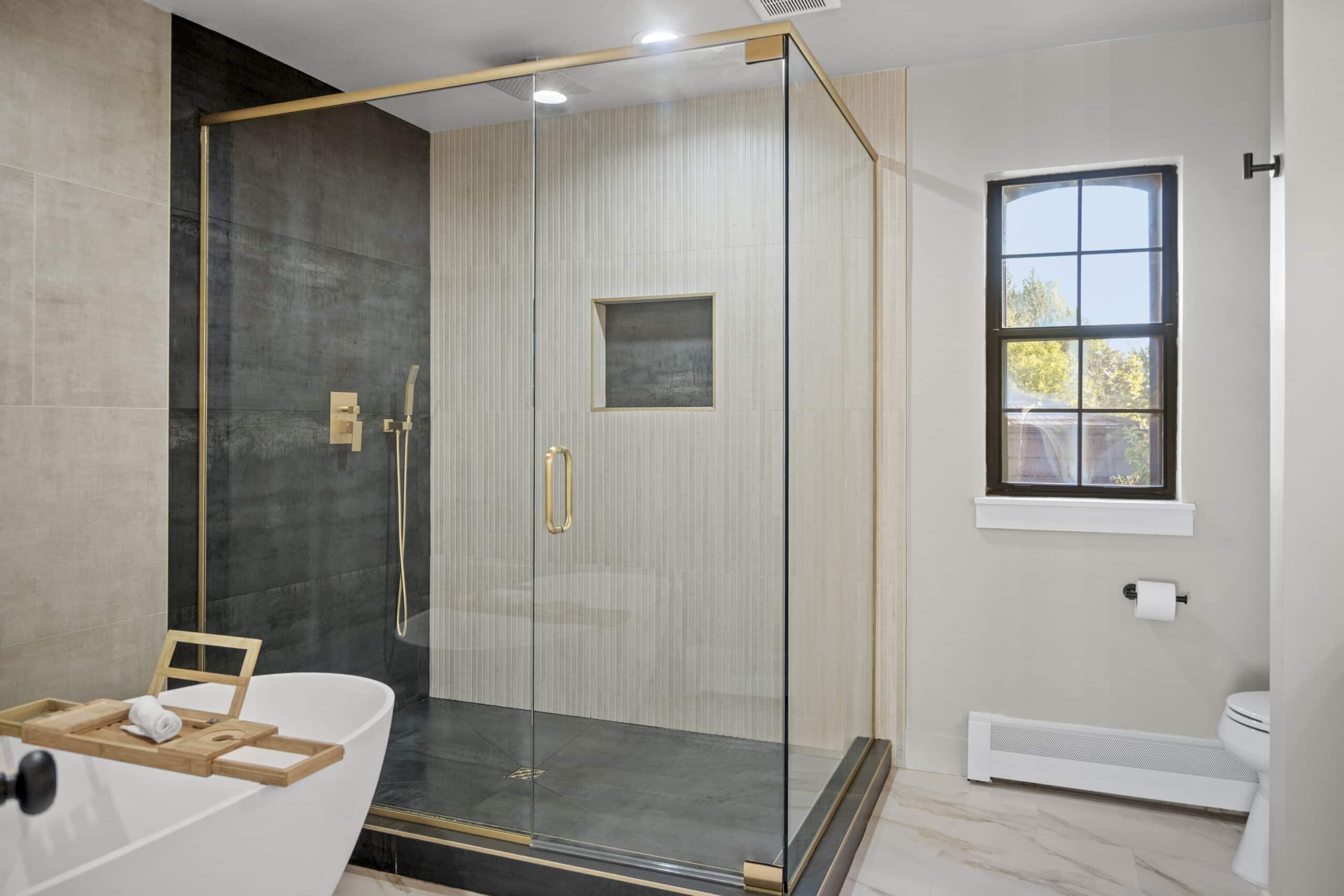 Elegant bathroom featuring a freestanding tub, glass shower with gold fixtures, and large window for natural light.