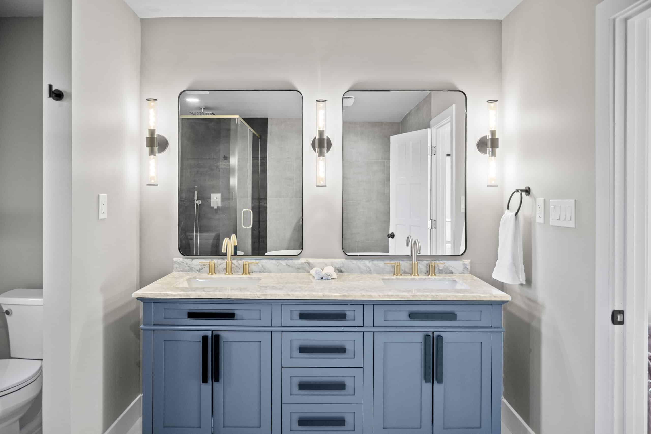 Remodeled bathroom with blue dual-sink vanity, marble countertop, gold fixtures, and tile shower.
