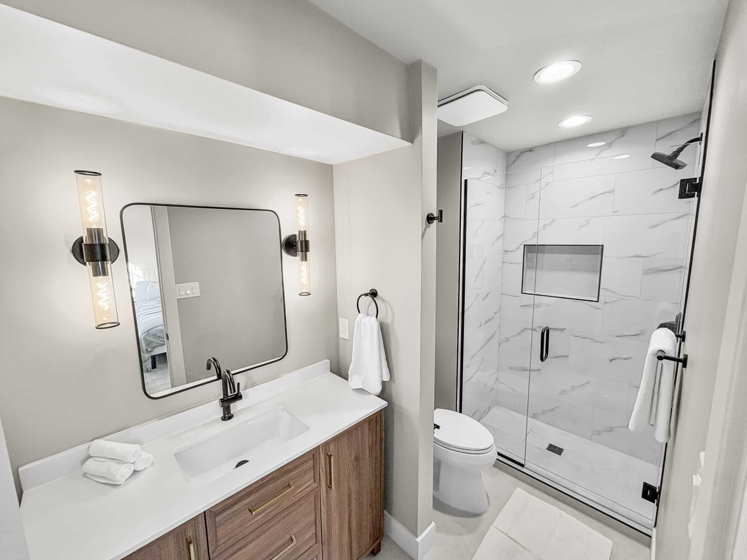 Remodeled bathroom with wood vanity, modern lighting, and glass shower with marble-style tile walls.